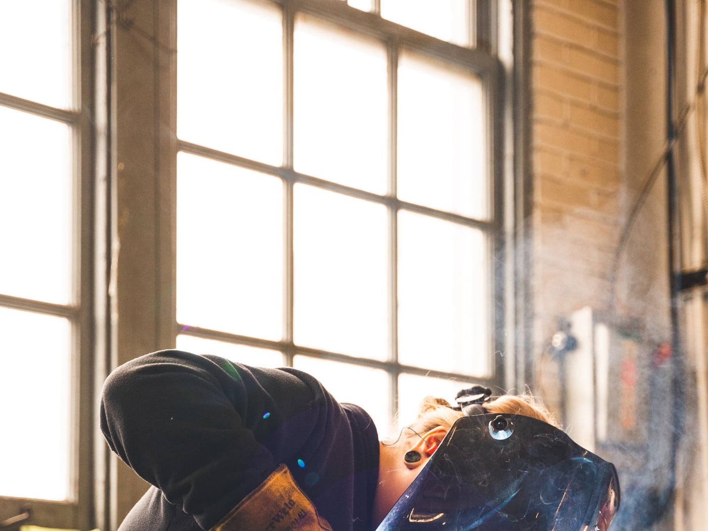 A welder in a helmet running a MIG gun along steel on a bench, lit by a tall shop window — representative imagery, not a specific Late Night Fabrication job