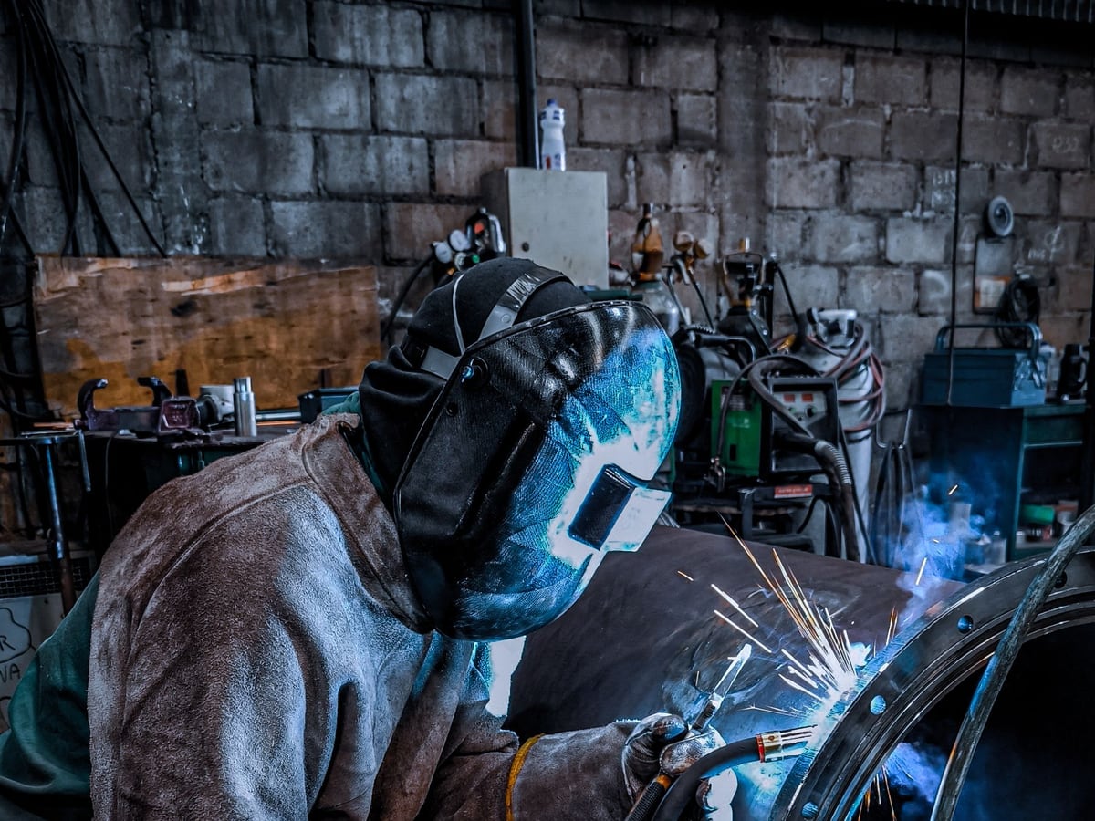 A welder in a helmet welding around a round steel section held in a fixture — representative imagery, not a specific Late Night Fabrication job
